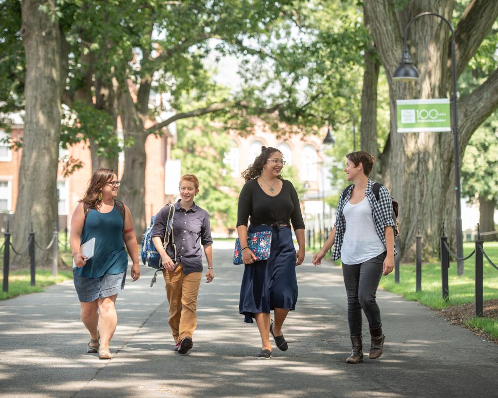 Four SSW students walk and talk down the path through the middle of campus, a paved walkway lined with black stanchions and trees, in front of a banner hanging from a lightpost that says "100 Years of Clinical Excellence"