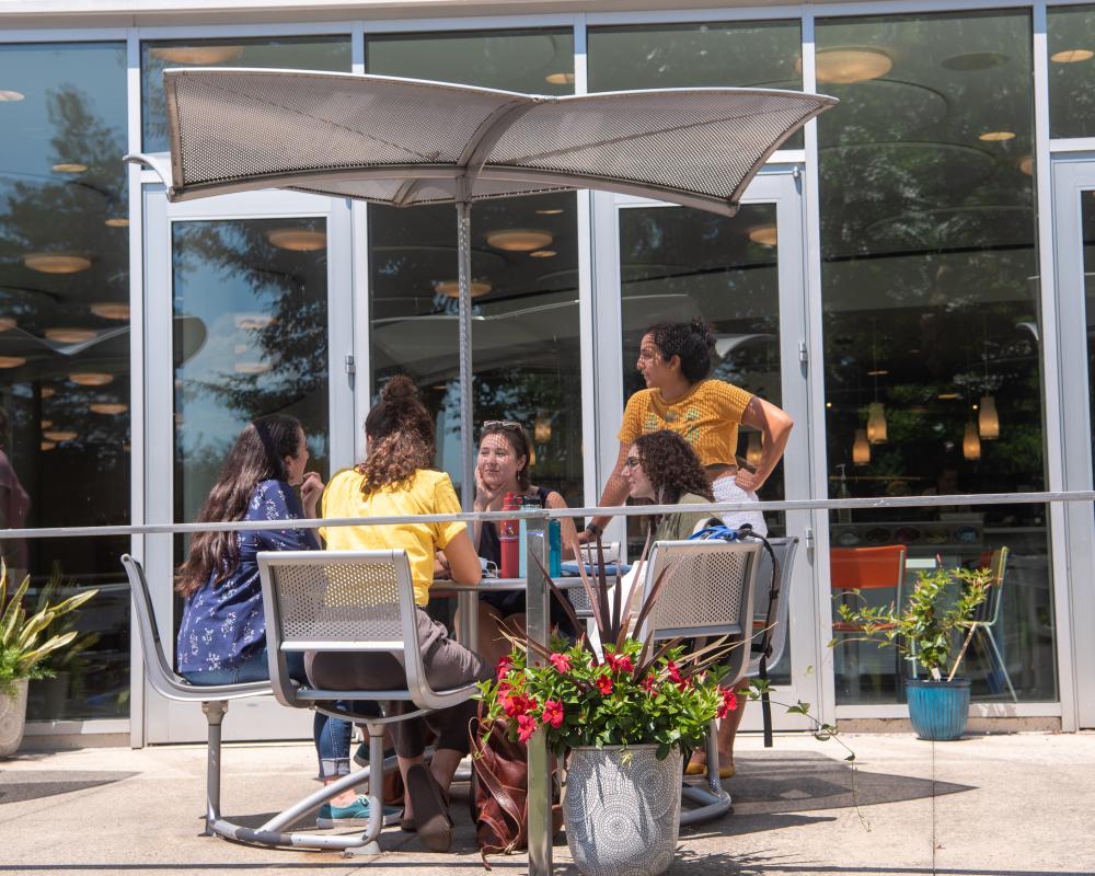 A group of students sits at a cafe table outside the Campus Center. They are talking and laughing. One student is standing and the rest are sitting. There are flowers in planters and large windows in the background.
