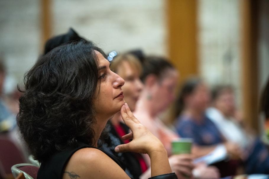 An audience member with shoulder-length dark hair holds two fingers to their chin as they look to the front of the room and listen in thoughtfulness