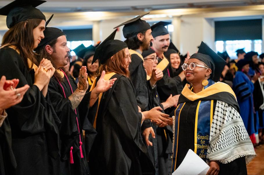 Wanja Kuria wearing black academic robes and cap and black and white kuffiyeh walking past other grads who are doing a standing ovation after her speech