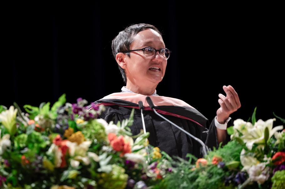 Miriam Yeung wearing black academic robes speaking behind flowers and greenery with small mic