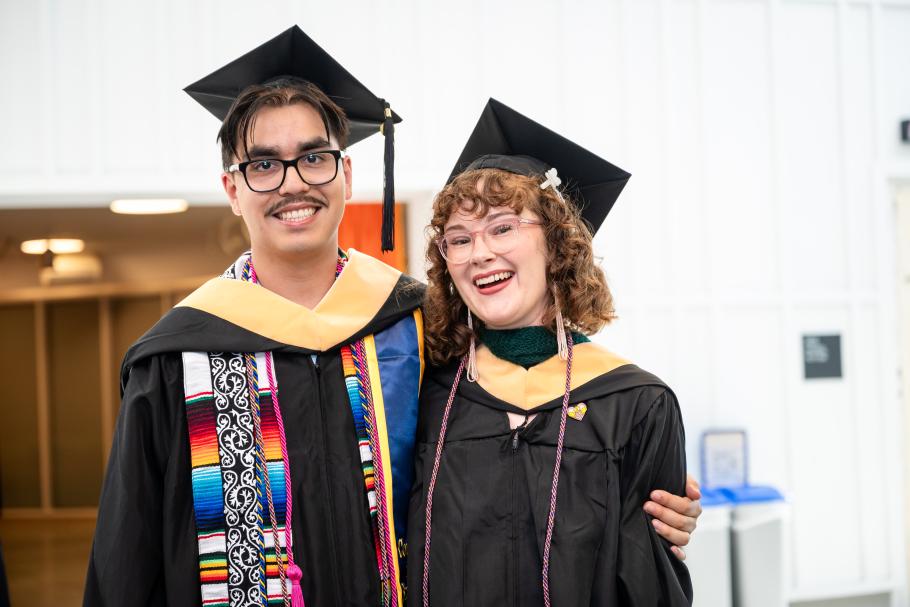 Two students wearing black academic robes, caps and yellow master's hoods smile and hug