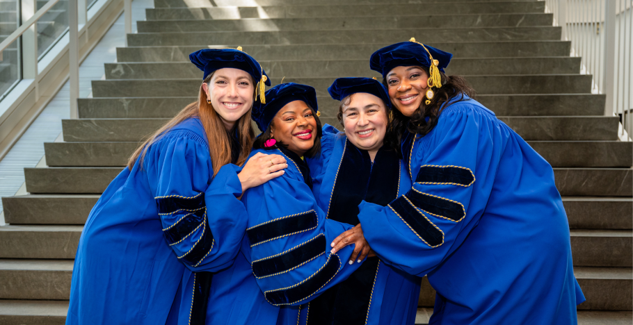 Four phd graduates in blue academic robes and tams hug and smile in front of cement stairs