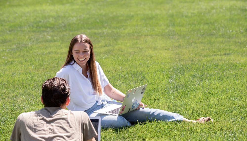 Molly Vencel sits on the grass talking with another student with a laptop open on her lap. 