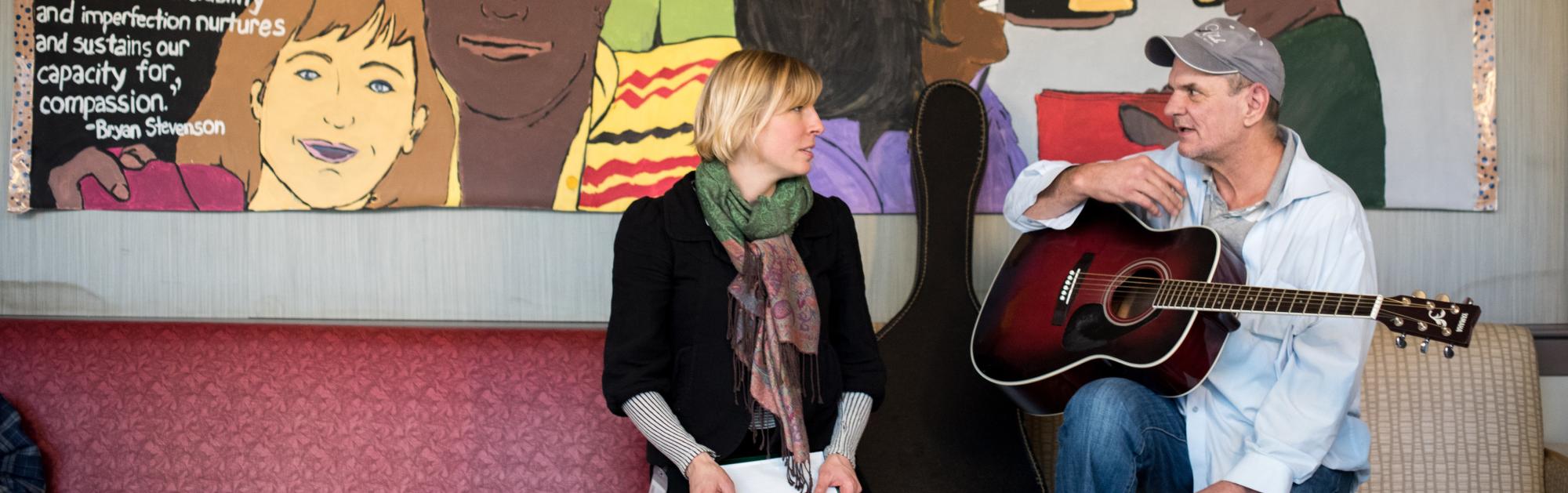 Two people sit on a bench in front of a large mural. One of them holds a notepad and the other holds a guitar. 