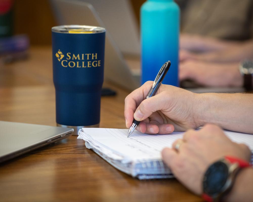 Photo of a person's hands writing in a notebook. A Smith College travel mug is in the background.