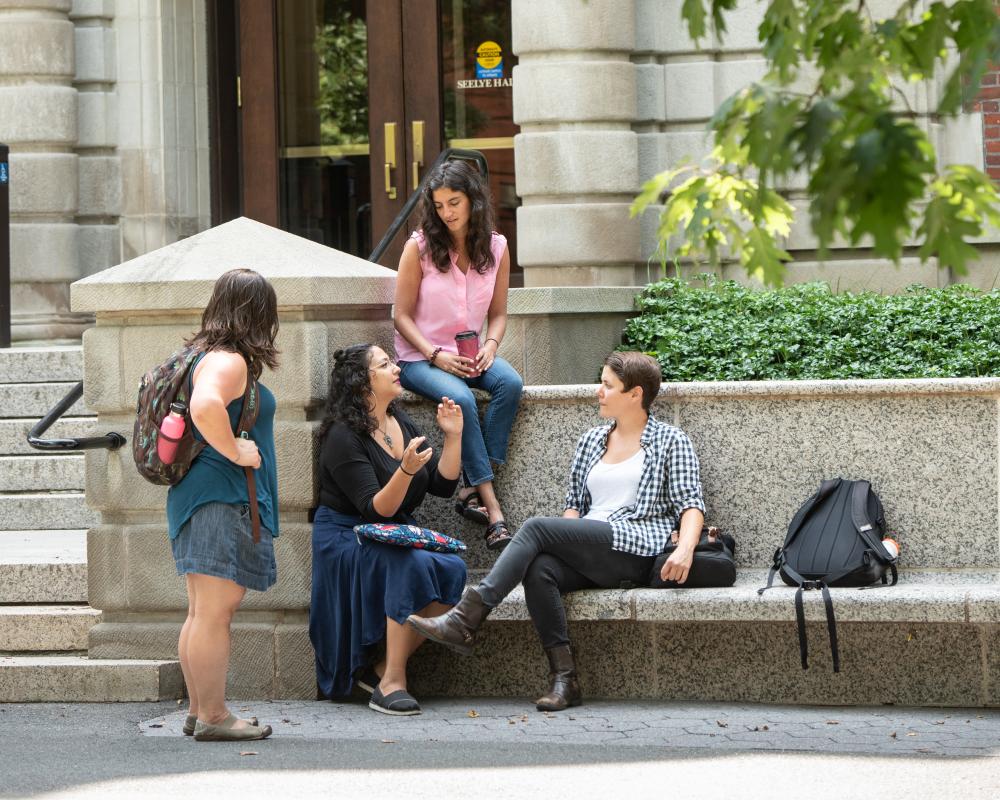 Four SSW students sit on stone benches outside the entrance of a building with stonework named "Seeleye Hall" during a bright day.