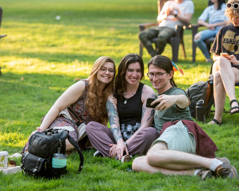 Three students sit on a sunny lawn taking a selfie.