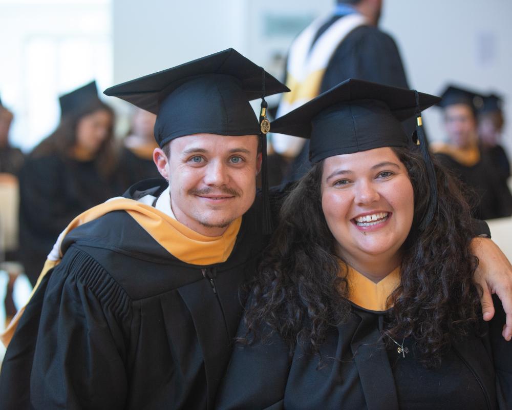 Two students wearing caps and gowns smile at the camera. 