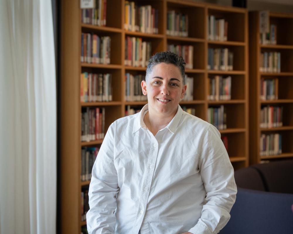 Lev Fein wearing white button down stands behind a wooden table with bookshelf behind in Neilson Library