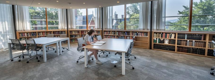 A student sits working at a table with rows of low bookshelves behind them. There is a second table in the room that is empty.