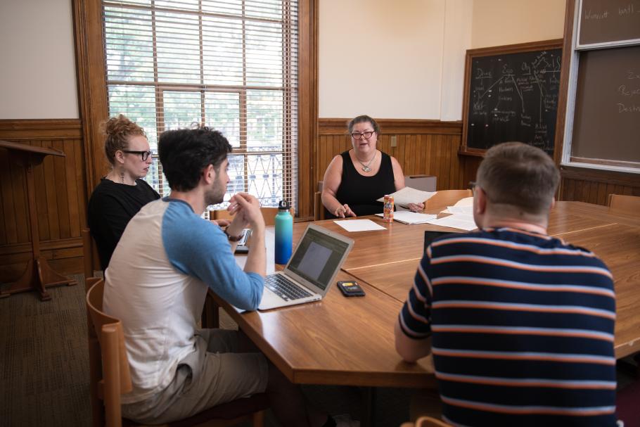 Four people sit around a table in a classroom engaged in discussion. There are laptops, cell phones and water bottles on the table. 