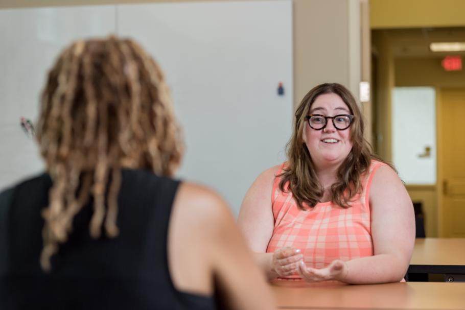 A person in a sleeveless plaid peach top with long hair and glasses engages in conversation in a classroom with a person with medium length brown and black hair and a black sleeveless top.