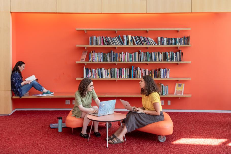 Three students sit in an orange study space in the Campus Center. Two are working together while the third reads in the background. There are open shelves filled with books lining the walls. 