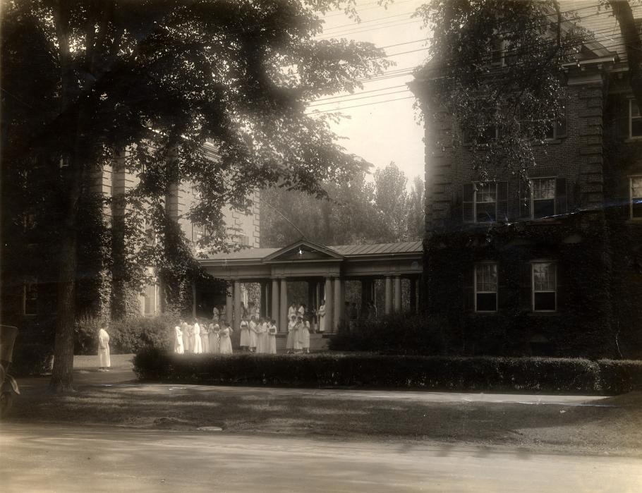 Black and white photo of Smith College campus from the early 1900s. There are brick buildings covered with ivy, a covered porch is in view with many people wearing white nursing clothing on it. 