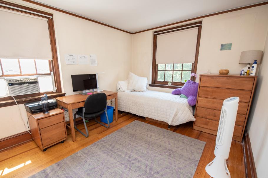 A dorm room with a twin bed, dresser, desk, small side table and a fan. There is a desktop computer on the desk. 