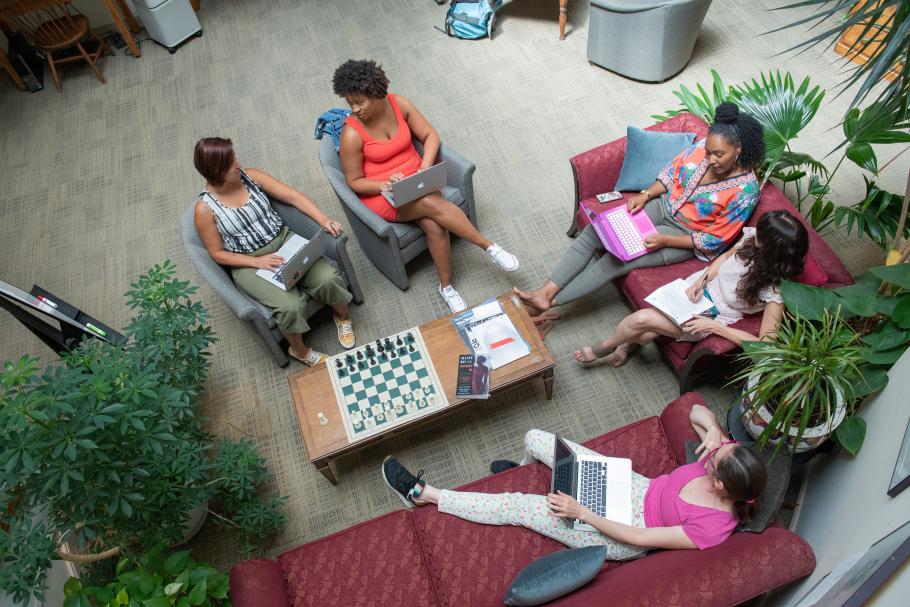 An overhead photo of five students sitting on couches and comfortable chairs with a coffee table in the center covered with various items and several lush green plants.