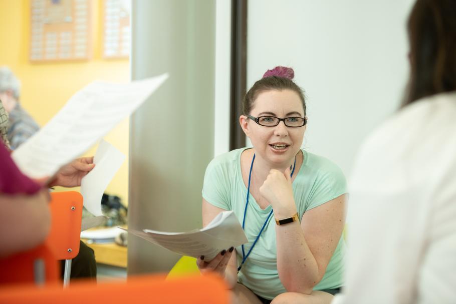 A professional education participant with a pink bun and mint shirt holds a paper packet, speaking at another participant.