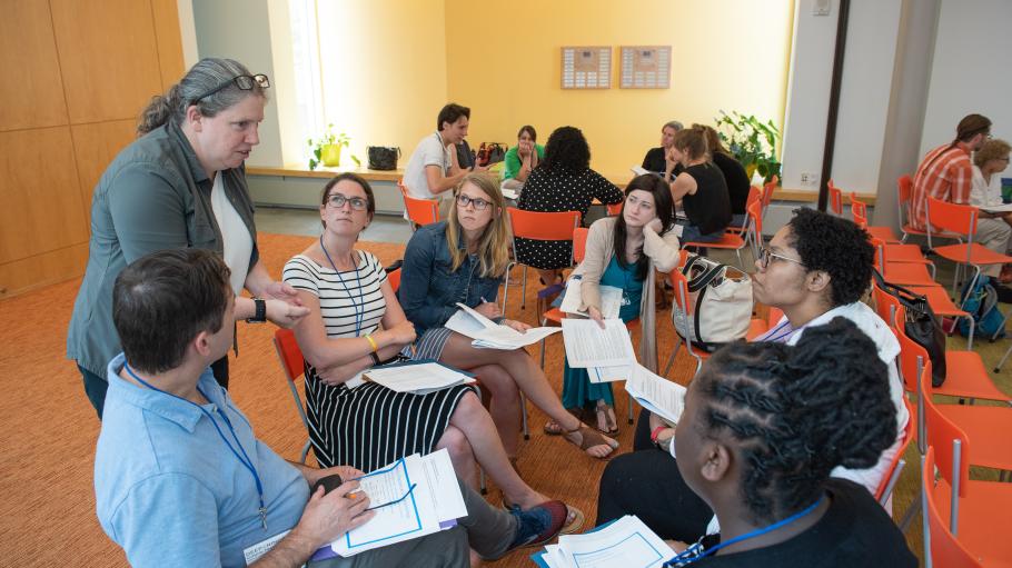 In a room with yellow walls, orange carpet, and orange chairs, a professional education instructor stands and speaks above a circle of participants holding paper packets.