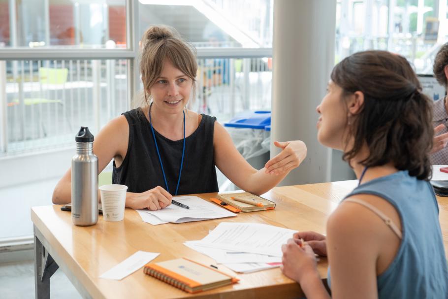 A professional education participant with brown hair in a bun on their head and a blank tank top motions to another participant sitting across the table with their head tilted.