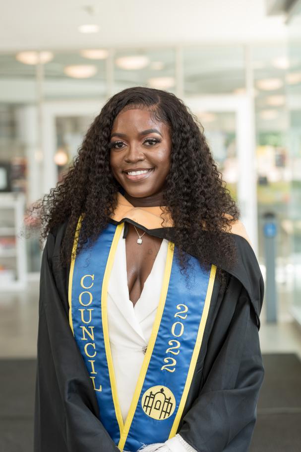 Beth Nanjala Luvisia wears a cap and gown over a white dress. A blue sash reading Council 2022 is draped across her shoulders.