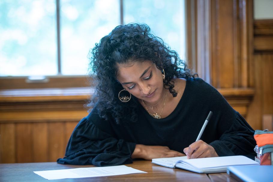 A student sits at a table writing in their notebook. They have long, dark, curly hair and are wearing a black top. 