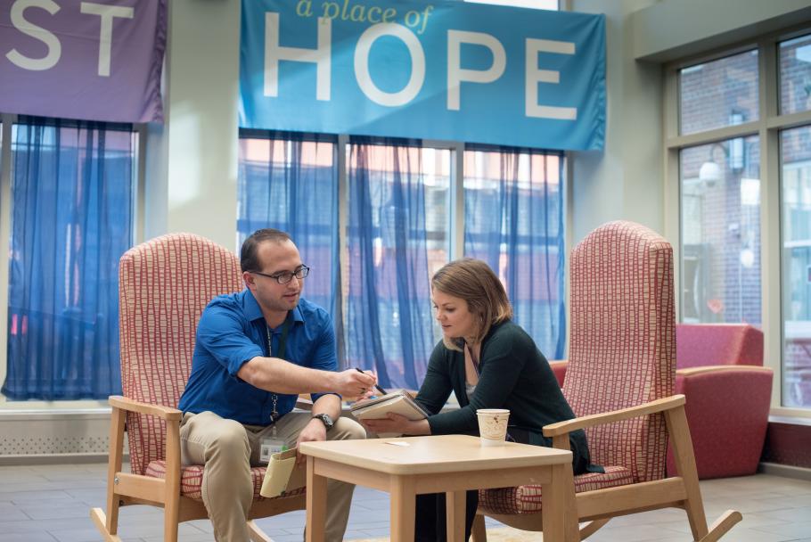 Two people sit in rocking chairs around a coffee table. They are looking at a clipboard. There is a banner hanging on the wall behind them that says hope. 