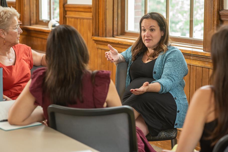 A faculty member sits in a classroom in conversation with three students.