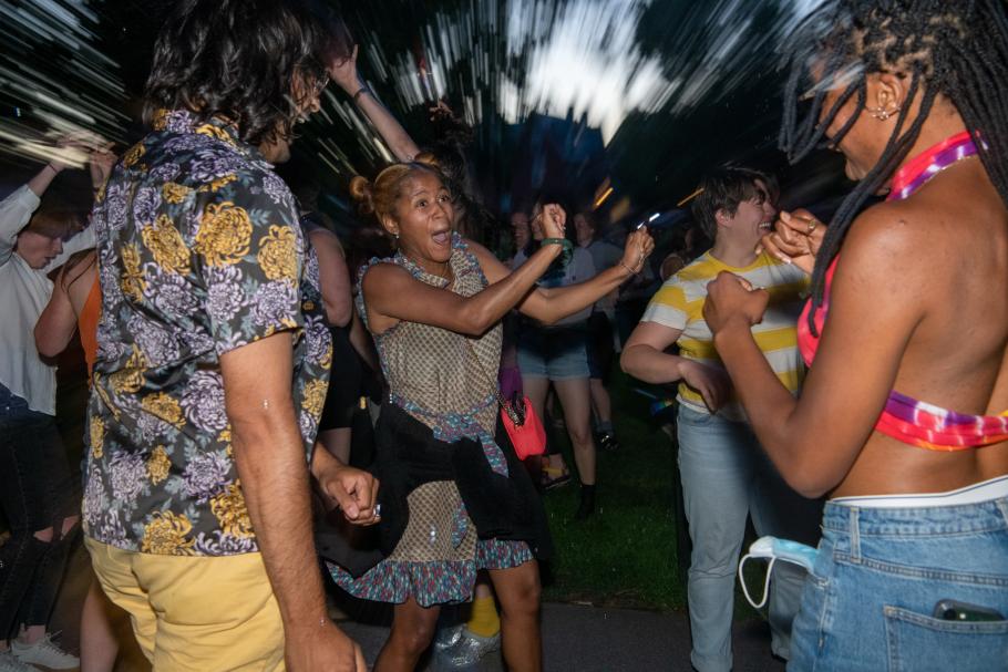 A group of students wearing summer attire dance outdoors in the darkness during a campus Pride celebration.