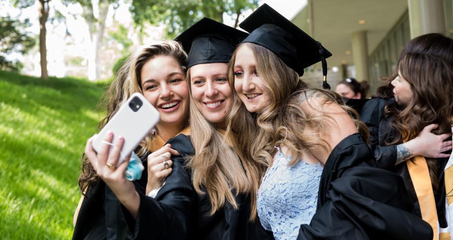 Three students standing outside the Campus Center wearing caps and gowns take a selfie before the 2022 commencement.