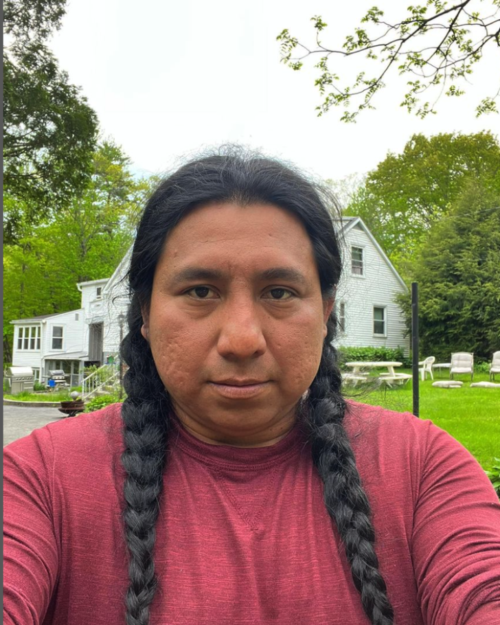 A person with long dark hair in two braids wearing a red shirt takes a selfie in front of a white house