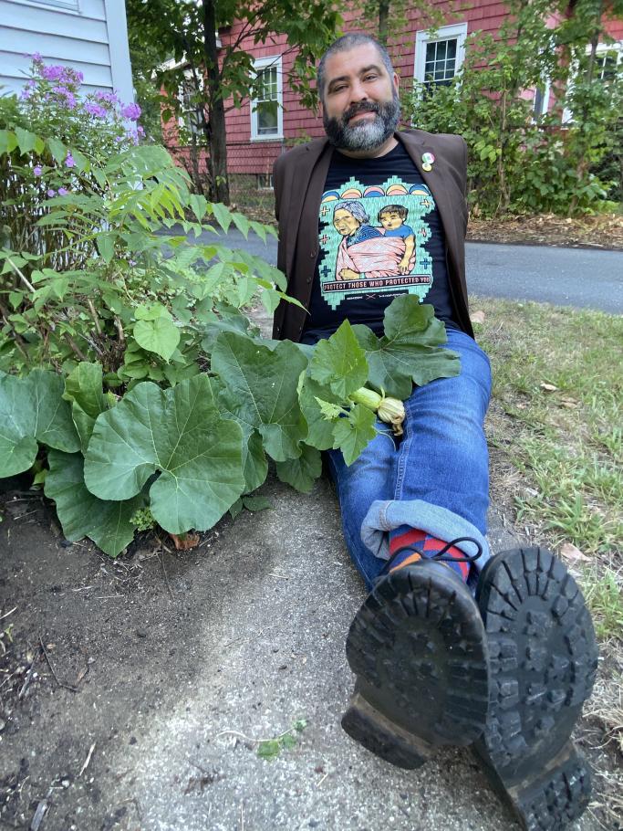 A person with short hair and a beard sits on the ground with a squash plant touching their legs, in jeans, with the soles of their shoes in the foreground