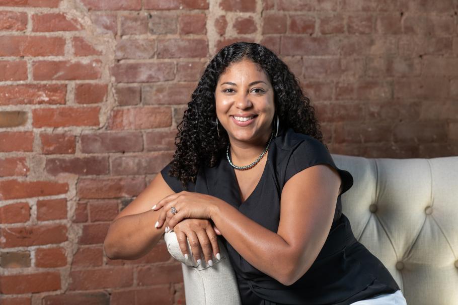 Jamie Daniels sits on a white couch in front of a brick wall. She is leaning on the arm of the couch and wearing a black top.