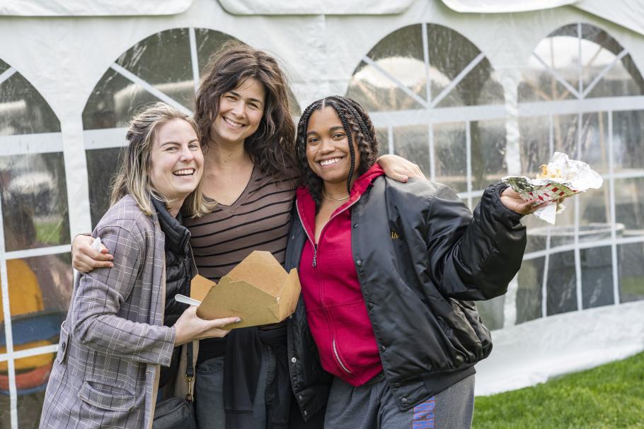 Three students smile during the Opening Ceremony. They are holding takeout containers and standing in front of a large white tent with see through window panels. 