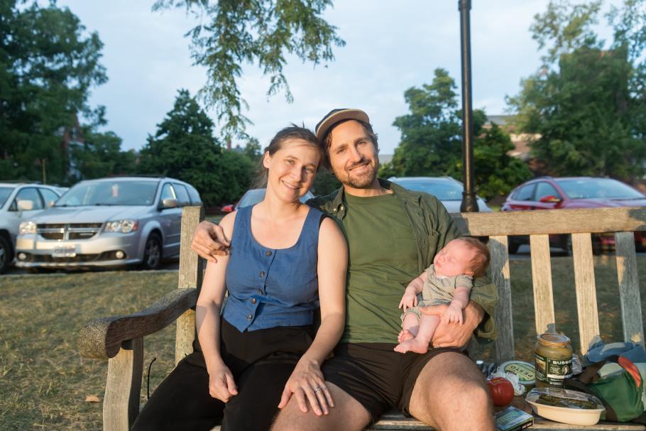 Two people sit on a bench in evening sunlight holding a baby.