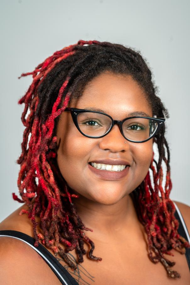 Zahria Thomas smiles at the camera with long red and black hair and glasses. 