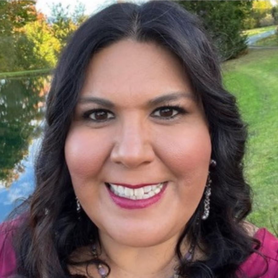 Luchanya Echeverria smiles close to the camera-with dark long hair and dark lipstick, trees and a pond and grass behind