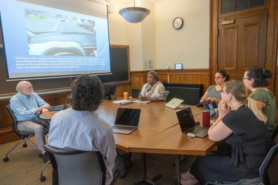 A group of Ph.D. students sit in the classroom engaged in conversation with one another. They are seated around a wooden table and a screen in the background displays an image. 