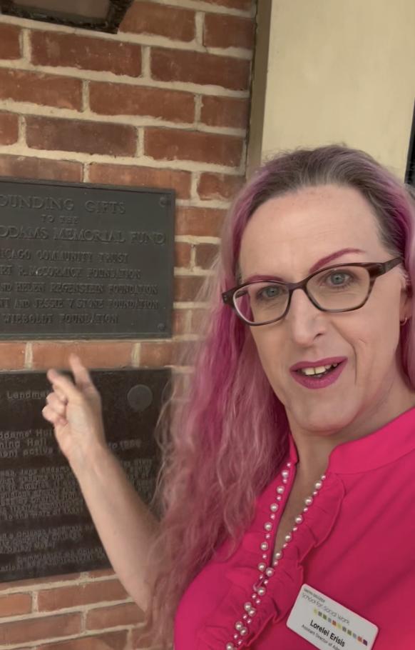 Lorelei Erisis stands in front of a brick wall at Hull-House pointing at a brass plaque 