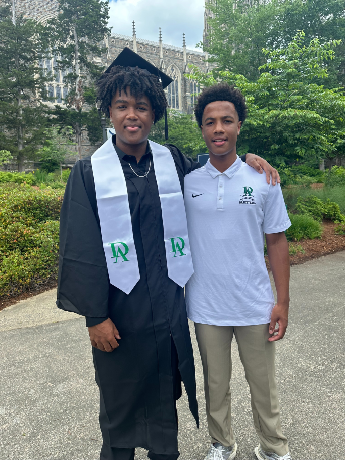 two people stand next to each other, one wearing academic robes and mortarboard and a white and green stole