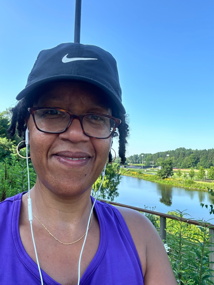 Jennifer Ashby Bullock takes a selfie wearing a baseball cap with headphones in. Over her shoulder is Paradise Pond, with green vegetation around