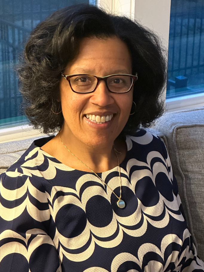 Jennifer Ashby Bullock headshot, sitting on grey couch wearing black glasses and black and white shirt