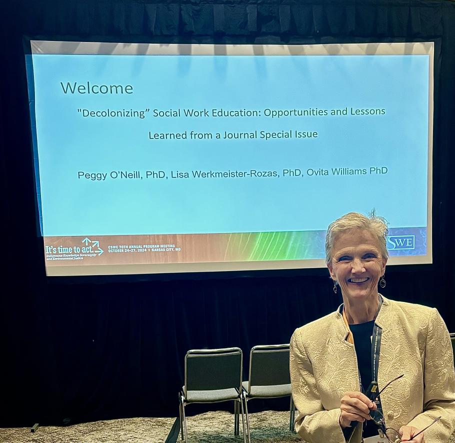 Peggy O'Neill sits in front of a screen that reads "Decolonizing" Social Work