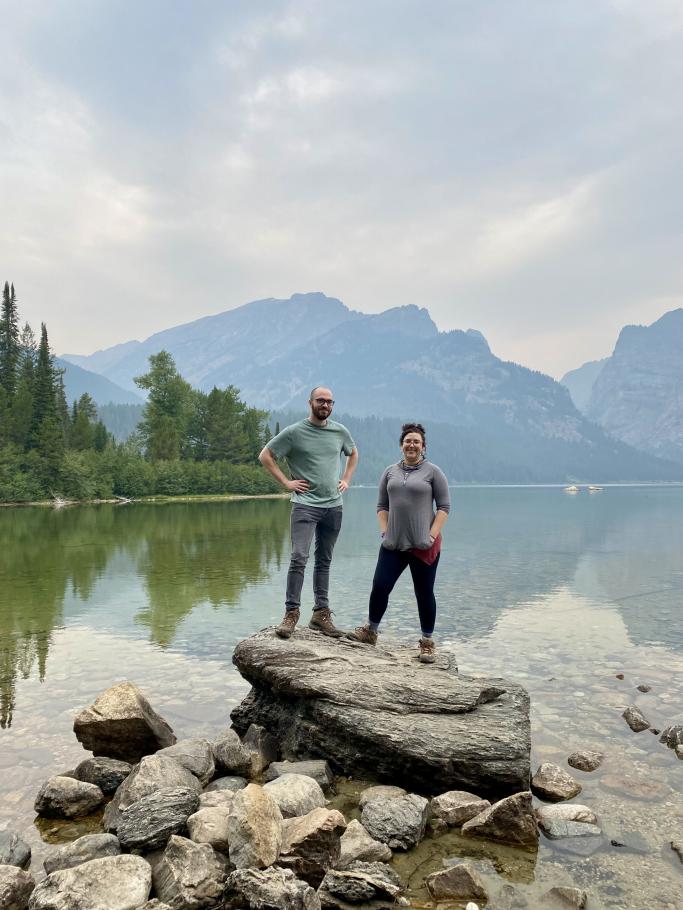 Hannah Biskind (right) and another person standing on a large rock at the shore of a large lake. Mountains behind