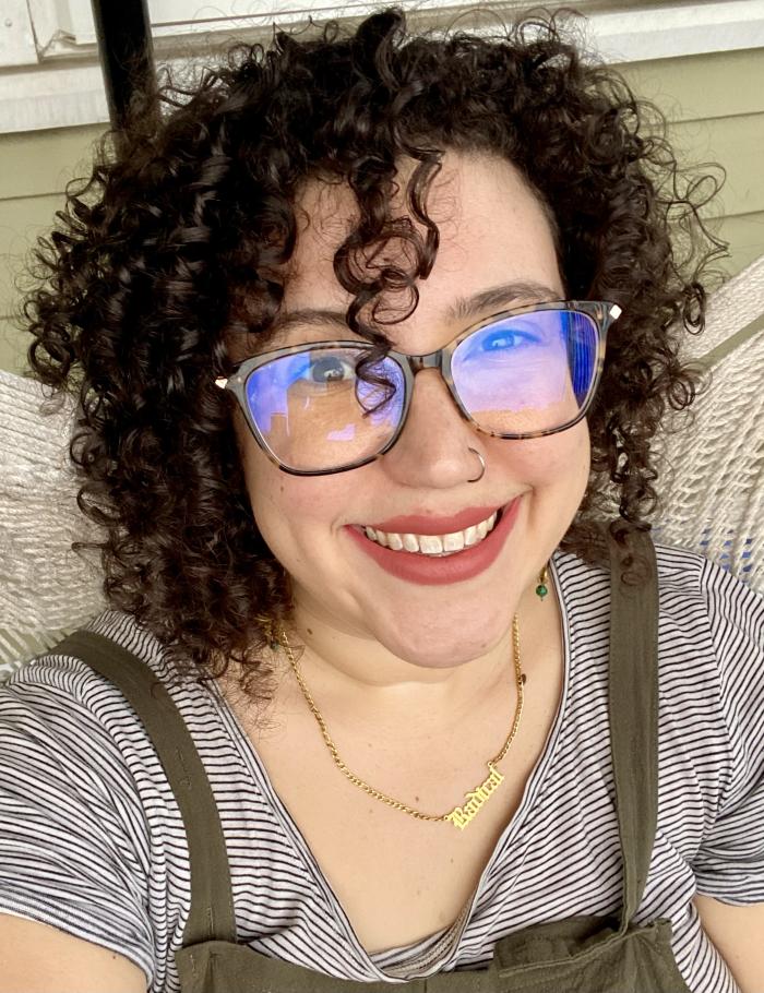 Selfie of Hannah Biskind, wearing glasses and a striped shirt sitting on a white wicker seat