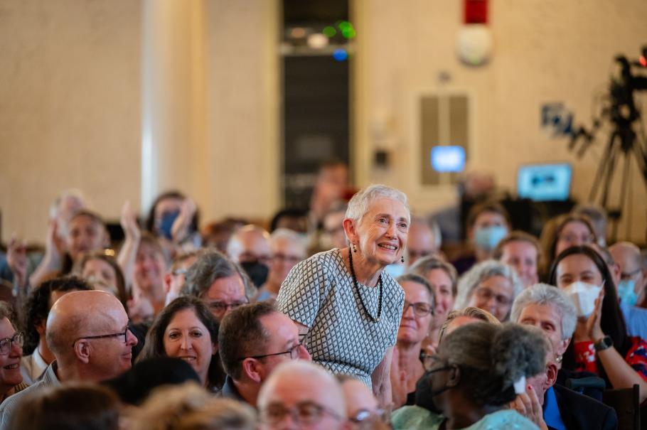 Sheila Buchdahl, a person standing up in a crowd, smiling. Walls behind are tan.