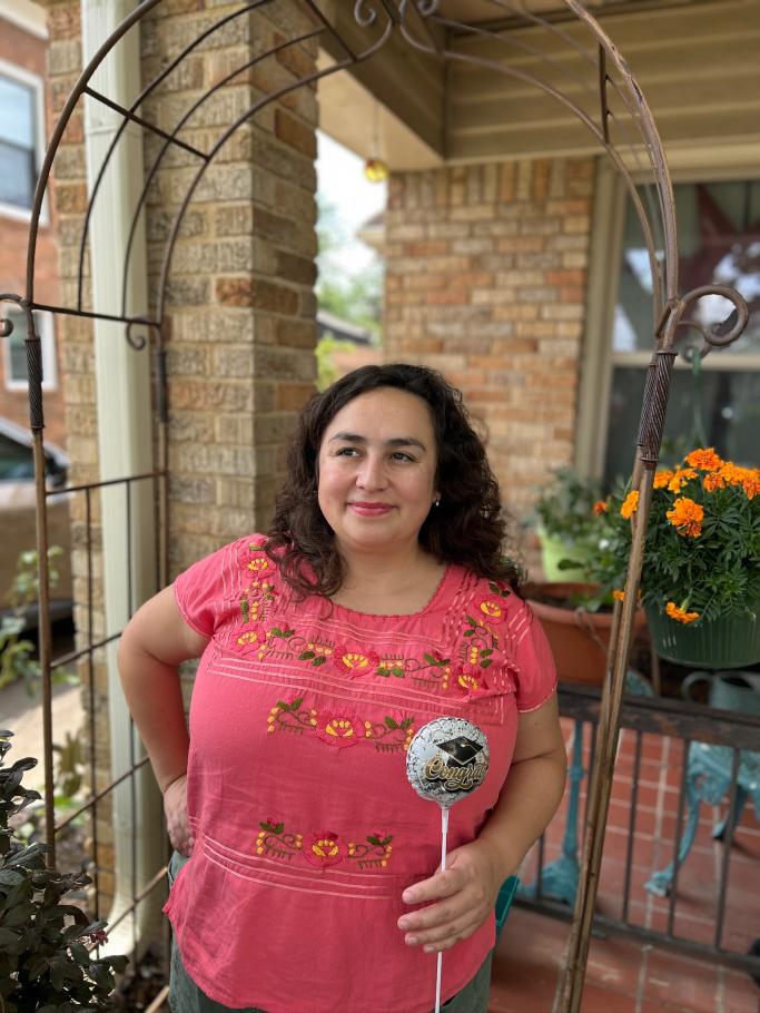 Maria Maldonado-Morales stands on a brick porch with arch behind, holding small balloon with graduation cap illustration