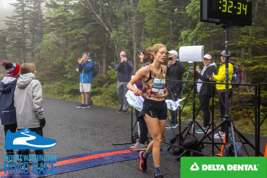 Rena Schwartz crossing a finish line running. People behind and metal stands.