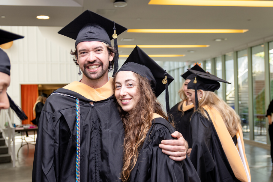 Iain Cooley, left, hugs another grad. Both wearing black academic robes and caps. Behind them are other grads, windows and ceiling lights