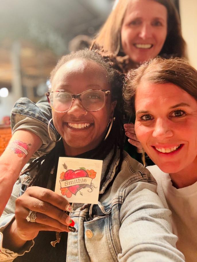 Janae Peters, Katie Potocnik Medina and Samantha Mishne hug, take selfie and Janae holds up a Smithie heart temporary tattoo